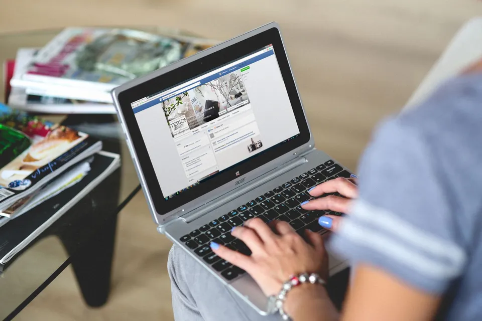 A person typing on a laptop beside a stack of magazines, working on website content