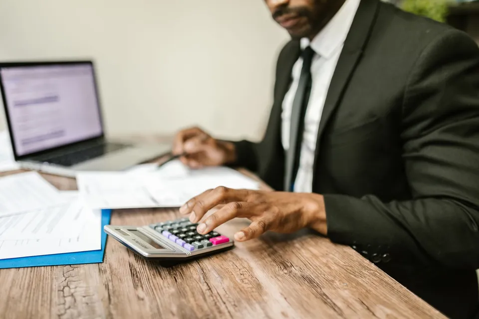 A person in a suit using a calculator over printed quote documents beside a laptop