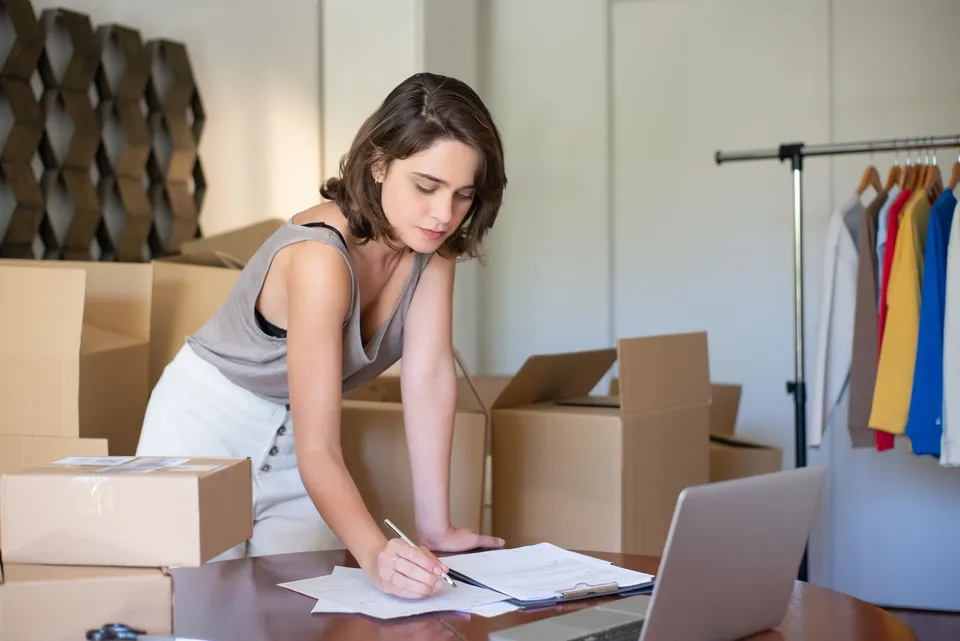 A store owner writing notes at a desk among shipping boxes with a clothing rack and laptop nearby